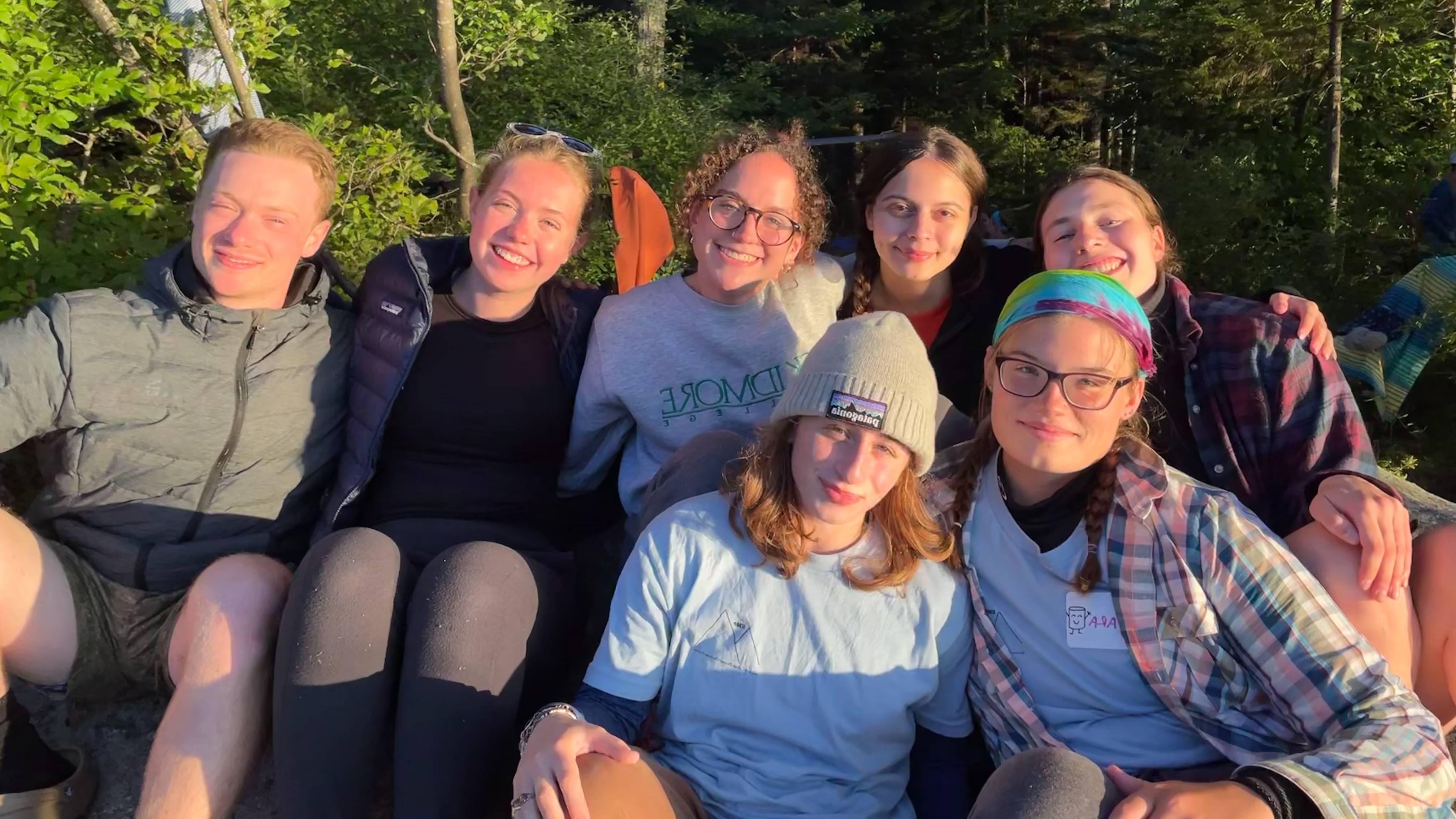 A group of seven students (including Catie Hamilton) sit and smile into the sun. Dense trees and greenery can be seen in the background.